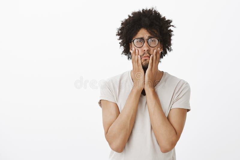 Sombere en verstoorde leuke mens met afrokapsel die, die wangen met handen drukken en met droevige en sombere uitdrukking staren stock foto Sombere en verstoorde leuke mens met afrokapsel die, die wangen met handen drukken en met droevige en sombere uitdrukking staren stock foto