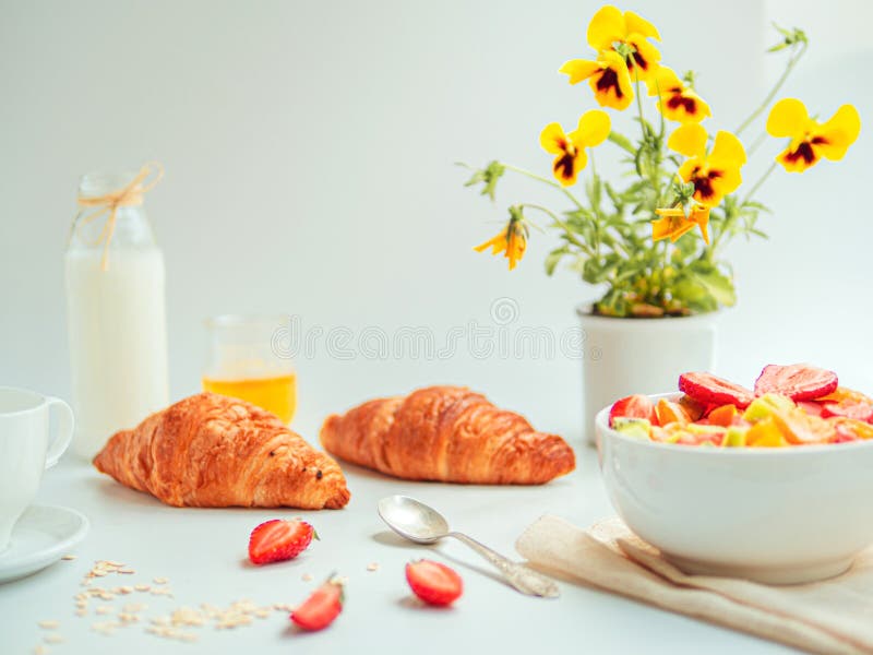Sommarfrukost med en bunke av sund sallad för ny frukt, giffel, och att mjölka Vit tabell med gula blommor royaltyfri bild Sommarfrukost med en bunke av sund sallad för ny frukt, giffel, och att mjölka Vit tabell med gula blommor royaltyfri bild