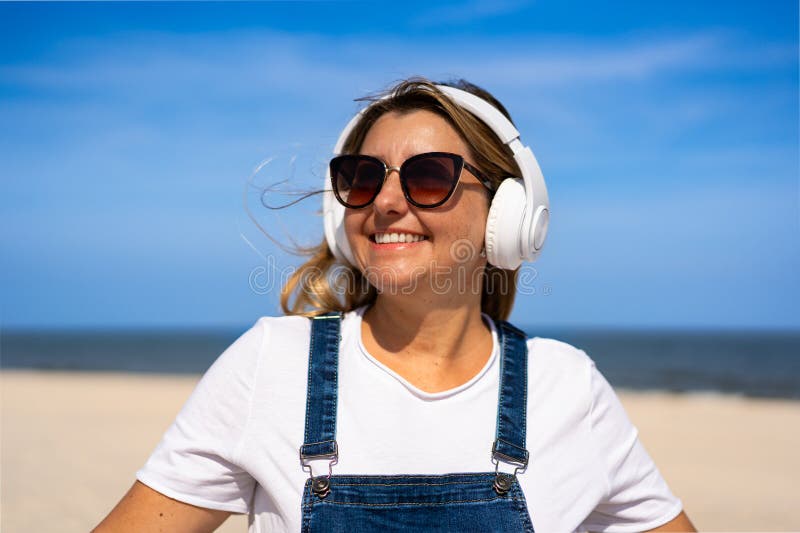 Sommartid med musik. Bild av en vacker, mogen kvinna som lyssnar på musik i hörlurar på sandstranden royaltyfri bild
