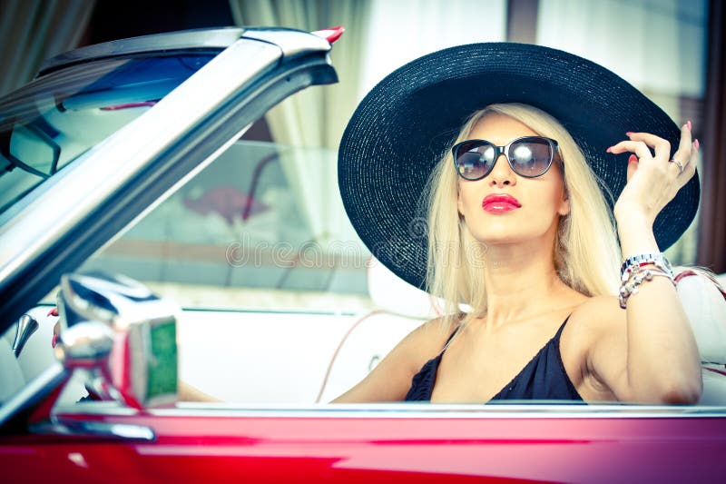 Outdoor summer portrait of stylish blonde vintage woman driving a convertible red retro car. Fashionable attractive fair hair female with black hat in red vehicle. Sunny bright colors, outdoors shot. Outdoor summer portrait of stylish blonde vintage woman driving a convertible red retro car. Fashionable attractive fair hair female with black hat in red vehicle. Sunny bright colors, outdoors shot.