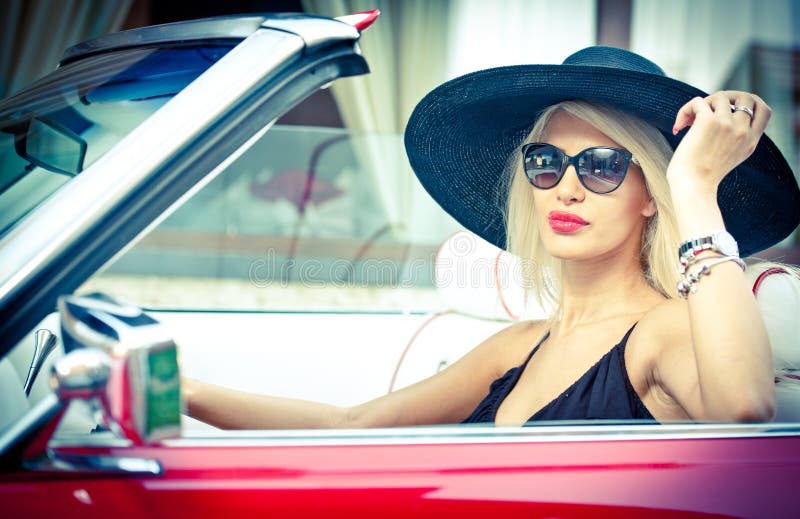 Outdoor summer portrait of stylish blonde vintage woman driving a convertible red retro car. Fashionable attractive fair hair female with black hat in red vehicle. Sunny bright colors, outdoors shot. Outdoor summer portrait of stylish blonde vintage woman driving a convertible red retro car. Fashionable attractive fair hair female with black hat in red vehicle. Sunny bright colors, outdoors shot.