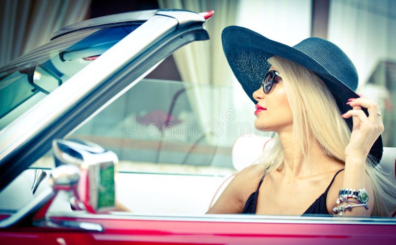 Outdoor summer portrait of stylish blonde vintage woman driving a convertible red retro car. Fashionable attractive fair hair female with black hat in red vehicle. Sunny bright colors, outdoors shot. Outdoor summer portrait of stylish blonde vintage woman driving a convertible red retro car. Fashionable attractive fair hair female with black hat in red vehicle. Sunny bright colors, outdoors shot.