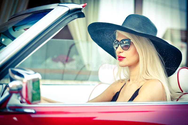 Outdoor summer portrait of stylish blonde vintage woman driving a convertible red retro car. Fashionable attractive fair hair female with black hat in red vehicle. Sunny bright colors, outdoors shot. Outdoor summer portrait of stylish blonde vintage woman driving a convertible red retro car. Fashionable attractive fair hair female with black hat in red vehicle. Sunny bright colors, outdoors shot.