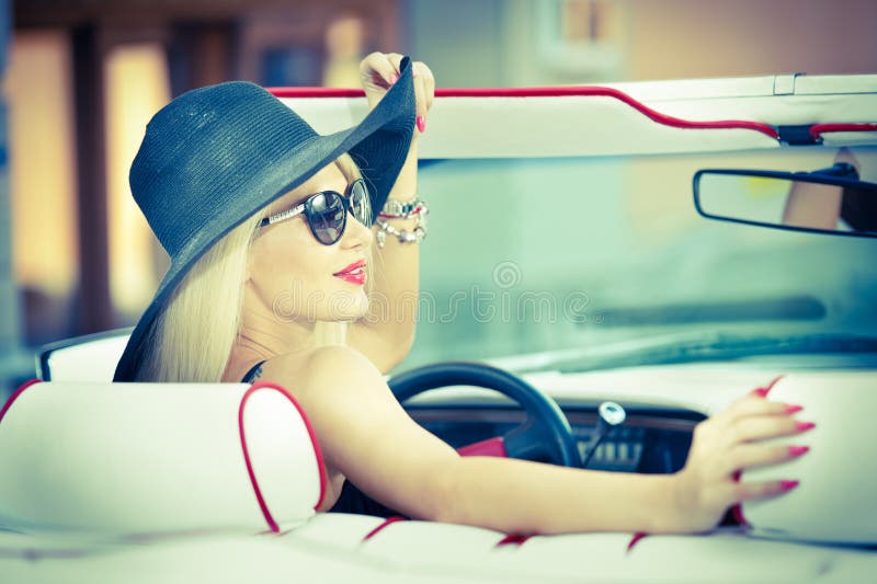 Outdoor summer portrait of stylish blonde vintage woman driving a convertible red retro car. Fashionable attractive fair hair female with black hat in red vehicle. Sunny bright colors, outdoors shot. Outdoor summer portrait of stylish blonde vintage woman driving a convertible red retro car. Fashionable attractive fair hair female with black hat in red vehicle. Sunny bright colors, outdoors shot.