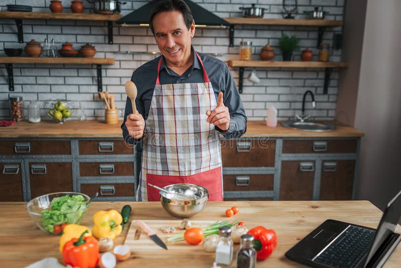 Cheerful man stand at table in kitchen and look on camera. He hold wooden spoon and show big thumb up. Laptop and colorful vegetables on table. Cheerful man stand at table in kitchen and look on camera. He hold wooden spoon and show big thumb up. Laptop and colorful vegetables on table