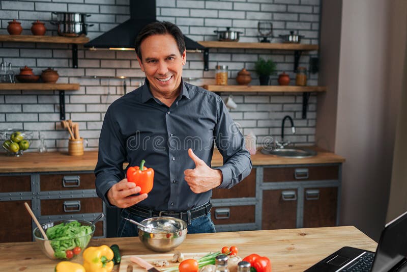 Positive man stand at table in kitchen and hold big thumb up. He look on camera and smile. Man hold pepper. Positive man stand at table in kitchen and hold big thumb up. He look on camera and smile. Man hold pepper