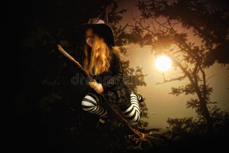 A little girl in traditional costume of a medieval witch and a pointed hat is flying on a broom through a dark forest. A little girl in traditional costume of a medieval witch and a pointed hat is flying on a broom through a dark forest