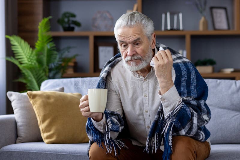 Sad senior gray-haired man sitting on sofa at home and suffering from illness. Covered with a blanket, holding a cup of hot drink and a napkin. Sad senior gray-haired man sitting on sofa at home and suffering from illness. Covered with a blanket, holding a cup of hot drink and a napkin.