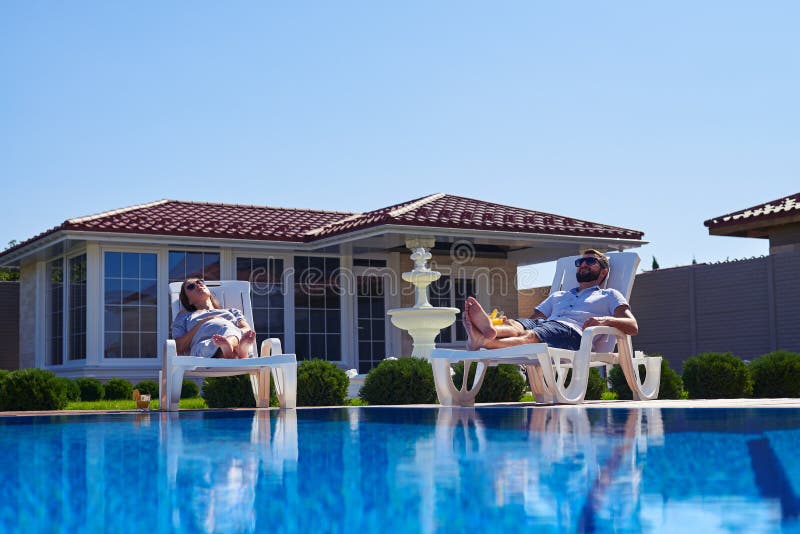 Sorglose Paare, die Sonnenbräune unter der Sonne nahe Pool erhalten lizenzfreies stockbild Sorglose Paare, die Sonnenbräune unter der Sonne nahe Pool erhalten lizenzfreies stockbild