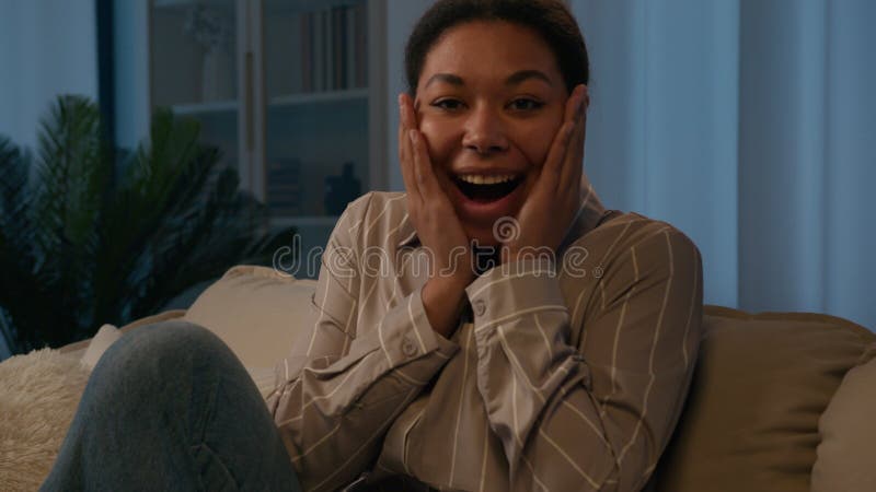 Sorprendida feliz mujer afroamericana de etnia hembra dueña en el sofá en la noche en casa mirando la cámara