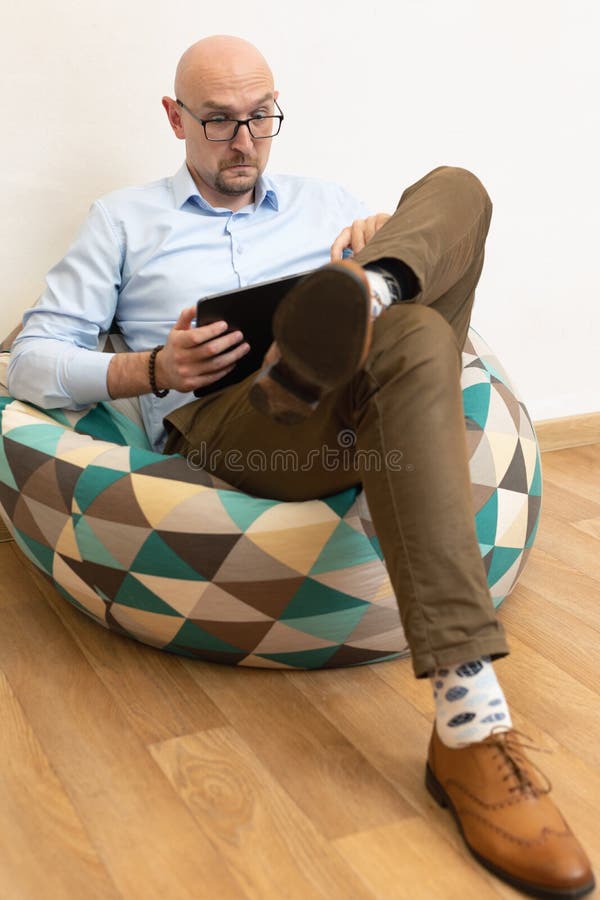 Sorprendido empresario calvo en gafas sosteniendo tableta digital mientras se sienta en la silla del bolso con mirada aturdida imagen de archivo libre de regalías