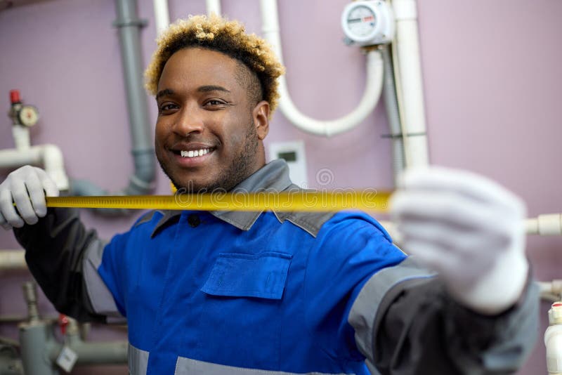 Professional worker in white gloves and overalls holds a tape measure in his hands. Black man with a toothy smile standing in the boiler room looks at the camera. Professional worker in white gloves and overalls holds a tape measure in his hands. Black man with a toothy smile standing in the boiler room looks at the camera