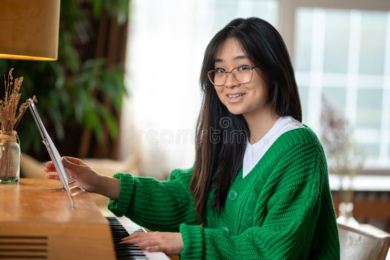 Sorridente garota asiática sentada perto do piano em casa foto de stock