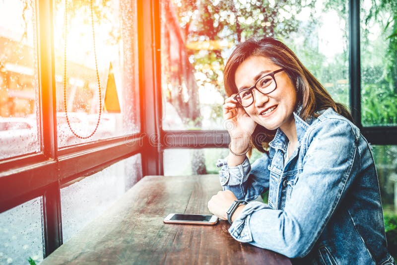 Sorriso asiatico di seduta della donna dei pantaloni a vita bassa del nerd di vetro in caffè delle finestre di vetro immagine stock libera da diritti Sorriso asiatico di seduta della donna dei pantaloni a vita bassa del nerd di vetro in caffè delle finestre di vetro immagine stock libera da diritti