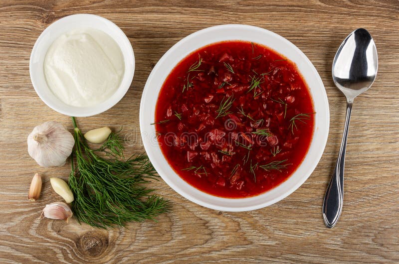 Sour cream in bowl, dill, garlic, plate with borscht, spoon on table. Top view