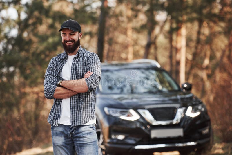 Sourire pour la caméra Homme barbu près de sa toute nouvelle voiture noire dans le concept de vacances en forêt photos libres de droits Sourire pour la caméra Homme barbu près de sa toute nouvelle voiture noire dans le concept de vacances en forêt photos libres de droits