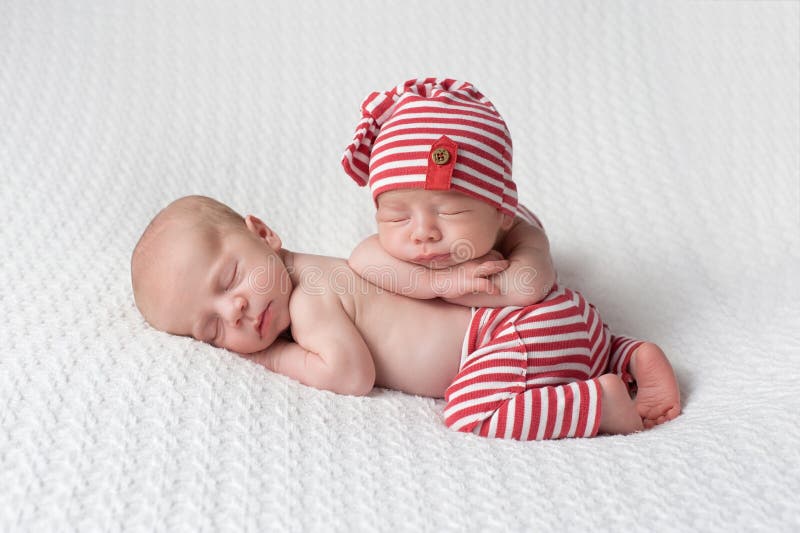 One month old fraternal, twin, newborn baby boys wearing red and white striped, up-cycled beanie and pants. They are stacked one on top of the other and sleeping on a white blanket. One month old fraternal, twin, newborn baby boys wearing red and white striped, up-cycled beanie and pants. They are stacked one on top of the other and sleeping on a white blanket.