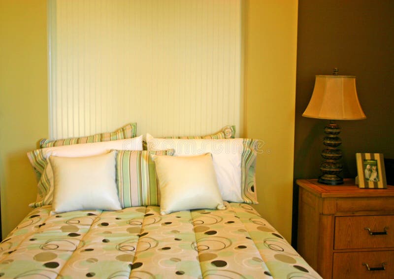 Modern bedroom with bedspread of circles, assortment of pillows, and lamp on bedside table, all against an unusual headboard and gold wall. Modern bedroom with bedspread of circles, assortment of pillows, and lamp on bedside table, all against an unusual headboard and gold wall