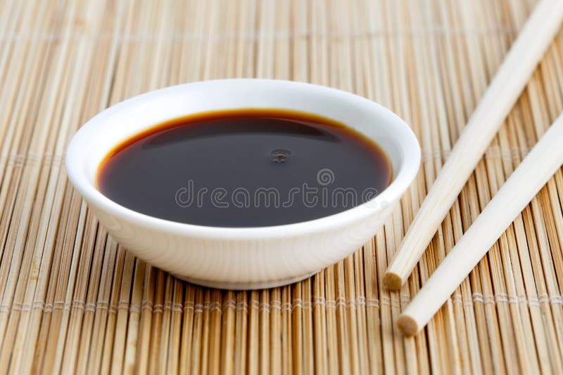 Soya sauce in white bowl on bamboo mat with wooden chopsticks.