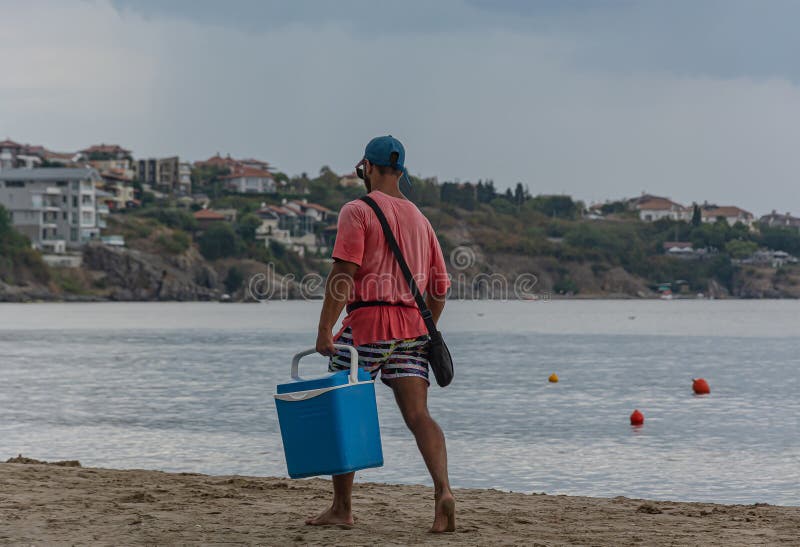 Sozopol, Bulgaria - 09/06/2018: a man in a red t-shirt with a blue box walks along the beach. Stock photo. Sozopol, Bulgaria - 09/06/2018: a man in a red t-shirt with a blue box walks along the beach. Stock photo