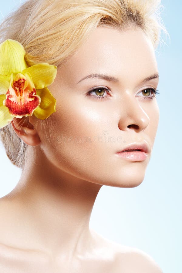 Healthcare. Spa. Wellness, beauty and skin care. Close-up portrait of beautiful woman with natural make-up, perfect shiny skin, yellow flower in her hair on blue sky background. Healthcare. Spa. Wellness, beauty and skin care. Close-up portrait of beautiful woman with natural make-up, perfect shiny skin, yellow flower in her hair on blue sky background