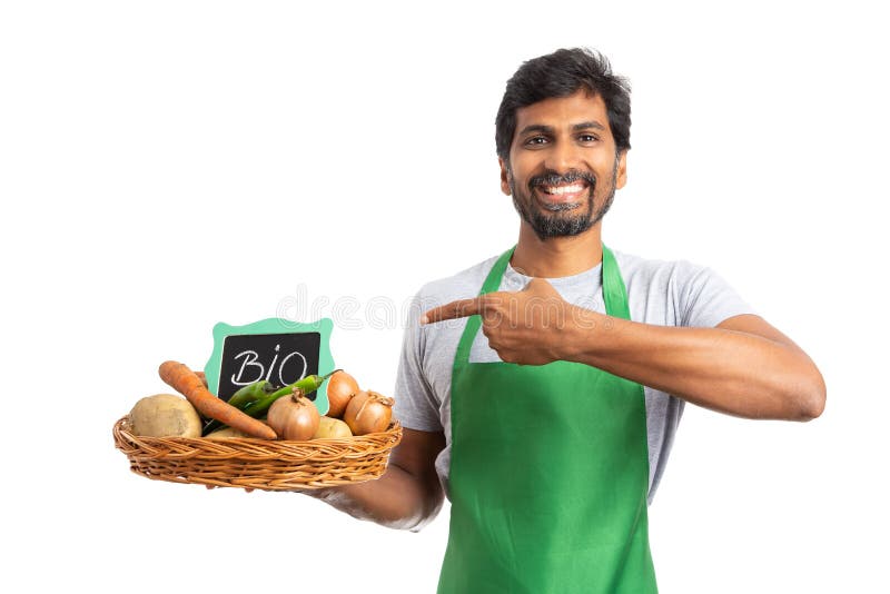 Indian male grocer smiling and presenting vegetable basket with bio sign using index finger isolated on white studio background as healthy lifestyle concept. Indian male grocer smiling and presenting vegetable basket with bio sign using index finger isolated on white studio background as healthy lifestyle concept