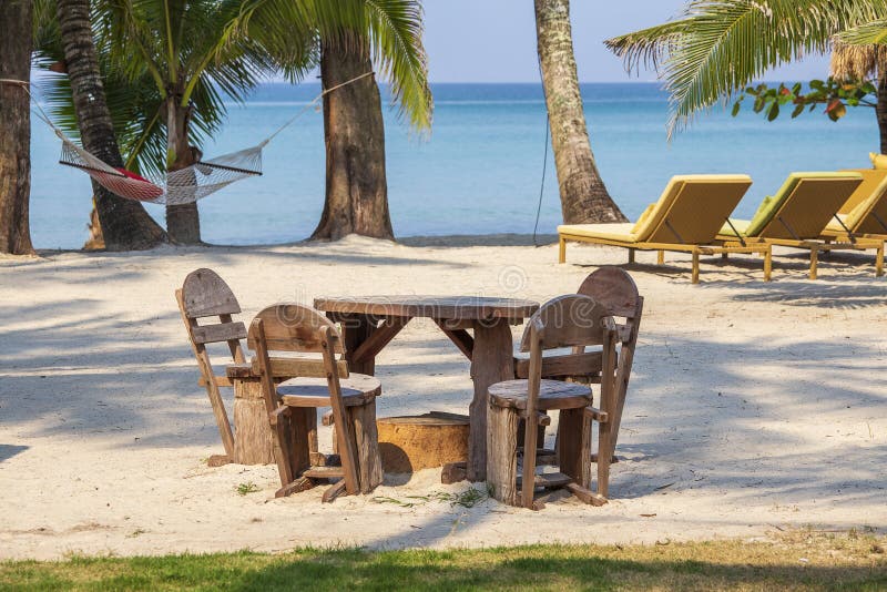 Spiaggia di sabbia tropicale con le palme di legno della tavola e delle sedie, delle chaise longue, dell'amaca e contro il contes fotografia stock Spiaggia di sabbia tropicale con le palme di legno della tavola e delle sedie, delle chaise longue, dell'amaca e contro il contes fotografia stock