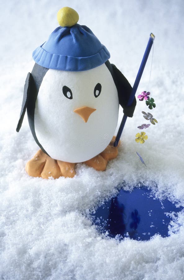 Large toy penguin holding fishing pole with sequins standing in snow before small blue pool. Penguin hand crafted by the photographer. Large toy penguin holding fishing pole with sequins standing in snow before small blue pool. Penguin hand crafted by the photographer