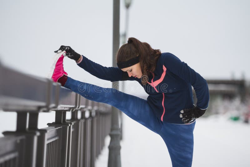 Sport concept - fitness girl model runner doing flexibility exercise for legs before run at snow winter promenade