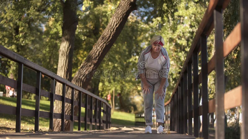 Sportieve hogere dame die haar knie houden die scherpe pijn voelen tijdens jogging, gezondheid stock afbeeldingen Sportieve hogere dame die haar knie houden die scherpe pijn voelen tijdens jogging, gezondheid stock afbeeldingen