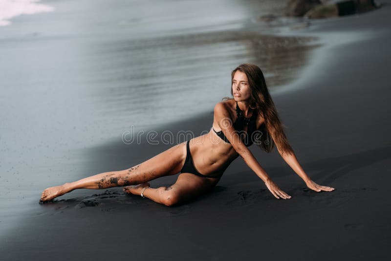 Sportig flicka med den sexiga kroppen i svart swimwear som vilar på den svarta sandstranden arkivbilder Sportig flicka med den sexiga kroppen i svart swimwear som vilar på den svarta sandstranden arkivbilder