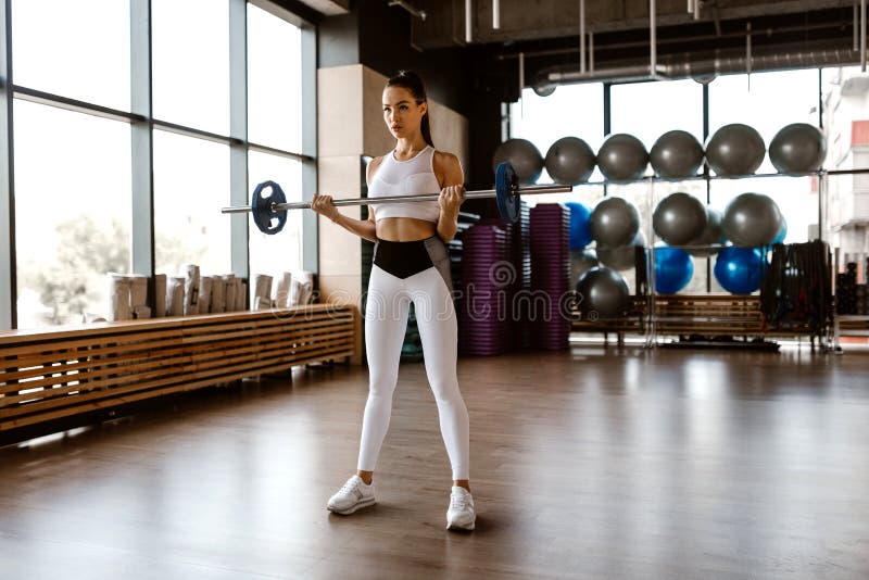 Sportowa dziewczyna ubierał w białych sportach wierzchołki i rajstopy dźwignięć barbell w gym zdjęcie stock Sportowa dziewczyna ubierał w białych sportach wierzchołki i rajstopy dźwignięć barbell w gym zdjęcie stock