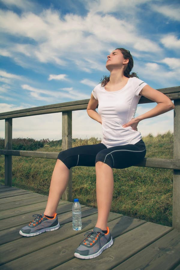 Athletic young woman in sportswear sitting touching her lower back muscles by painful injury, over a nature background. Sport injuries concept. Athletic young woman in sportswear sitting touching her lower back muscles by painful injury, over a nature background. Sport injuries concept.