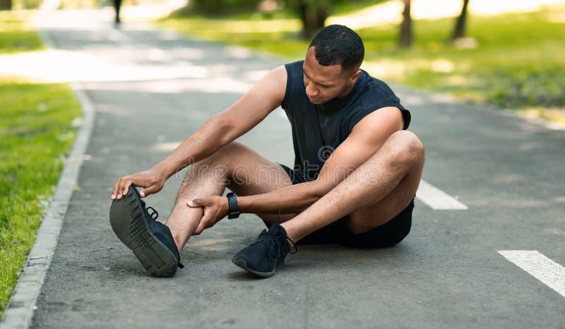 Sports injury. African American runner sitting on jogging track and feeling pain in his ankle, panorama