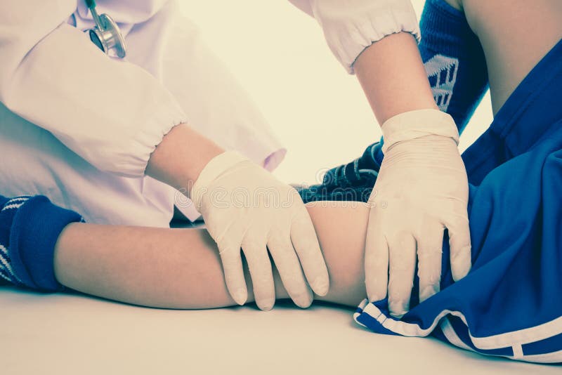 Sports injury. Youth soccer player in blue uniform with pain in knee. Doctor perform checking and first aid at knee trauma. Studio shot. Cream tones. Sports injury. Youth soccer player in blue uniform with pain in knee. Doctor perform checking and first aid at knee trauma. Studio shot. Cream tones.