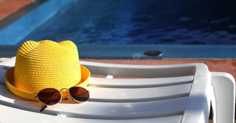 Sporttillbehör en gul hatt och solglasögon ligger på en vit däcksstol vid poolen. Solskydd för solskyddsmedel. semester royaltyfri foto