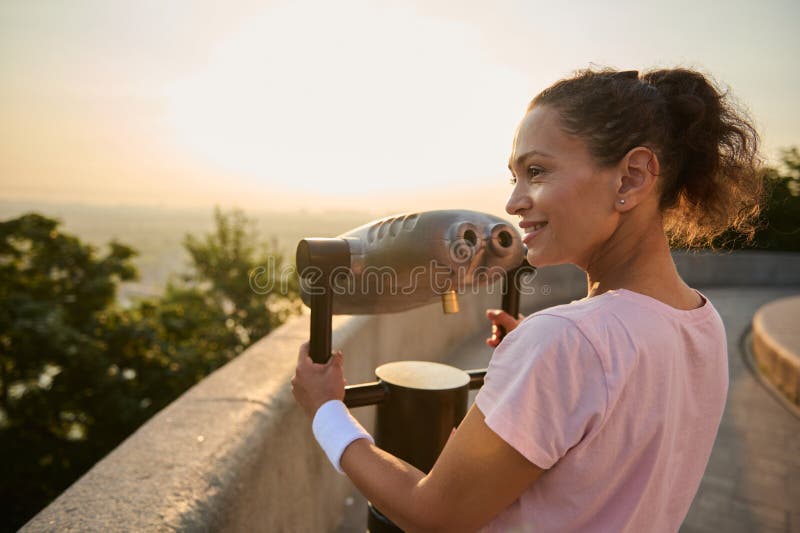 Sporty pretty woman looking through a telescope at the city. Athletic woman in a pink t-shirt is watching the beautiful green stock photos