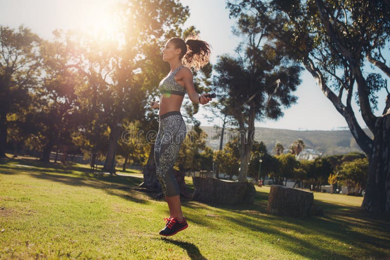 Side view of sporty woman skipping in a park on a sunny morning. Fit and athletic female exercising with a jump rope on the grass.