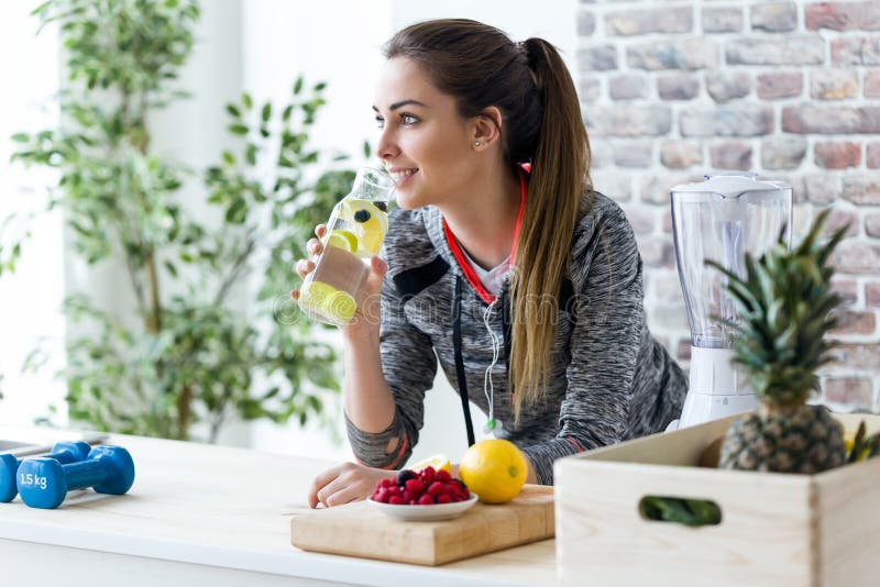 Sporty молодая женщина смотря косой пока выпивающ лимонный сок в кухне дома стоковая фотография rf Sporty молодая женщина смотря косой пока выпивающ лимонный сок в кухне дома стоковая фотография rf