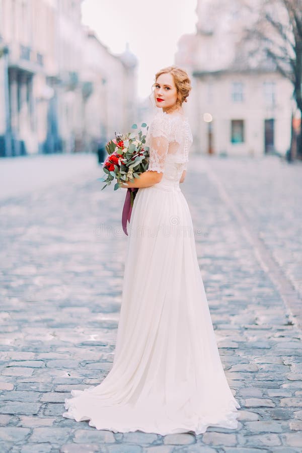 Sposa affascinante nel mazzo d'annata dal pizzo della tenuta lunga del vestito che esamina spalla nella macchina fotografica con immagini stock libere da diritti Sposa affascinante nel mazzo d'annata dal pizzo della tenuta lunga del vestito che esamina spalla nella macchina fotografica con immagini stock libere da diritti