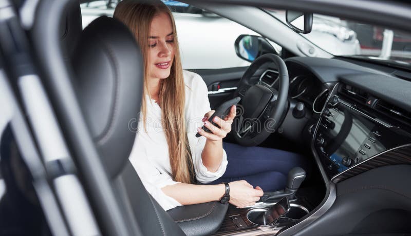 Sprawdzanie wiadomości w telefonie Piękna blondynka siedząca w nowym samochodzie z nowoczesnym czarnym wnętrzem zdjęcia stock Sprawdzanie wiadomości w telefonie Piękna blondynka siedząca w nowym samochodzie z nowoczesnym czarnym wnętrzem zdjęcia stock