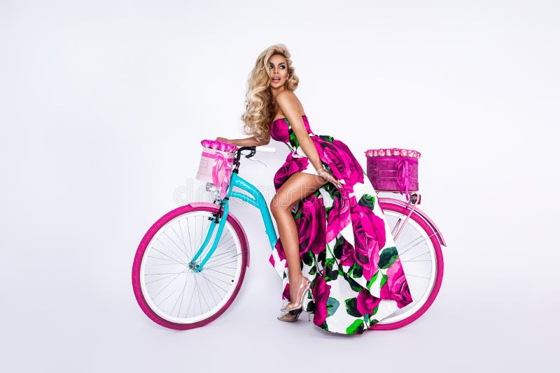 Spring fashion. Sexy blonde model in floral dress on colorful bike, decorated with flowers on white background. Spring concept.