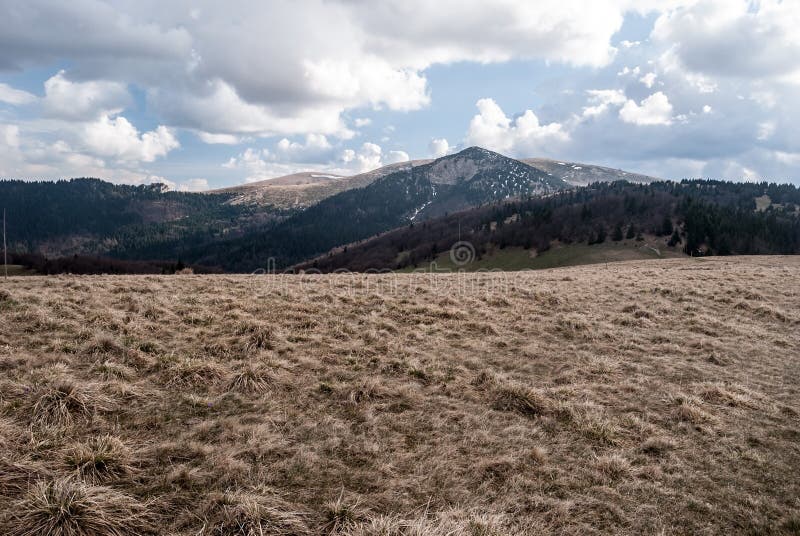 Jarné hory s horskou lúkou, kopce so snehovými poliami a modrá obloha s mrakmi