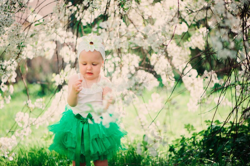 Spring portrait of cute baby girl in green skirt enjoying outdoor walk in blooming garden