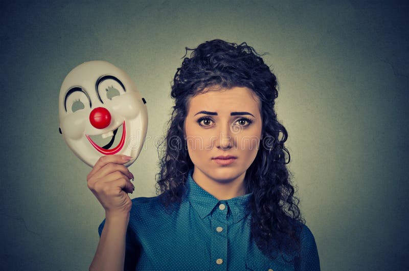 Portrait young upset worried woman with sad expression holding a clown mask expressing cheerfulness happiness on gray wall background. Portrait young upset worried woman with sad expression holding a clown mask expressing cheerfulness happiness on gray wall background