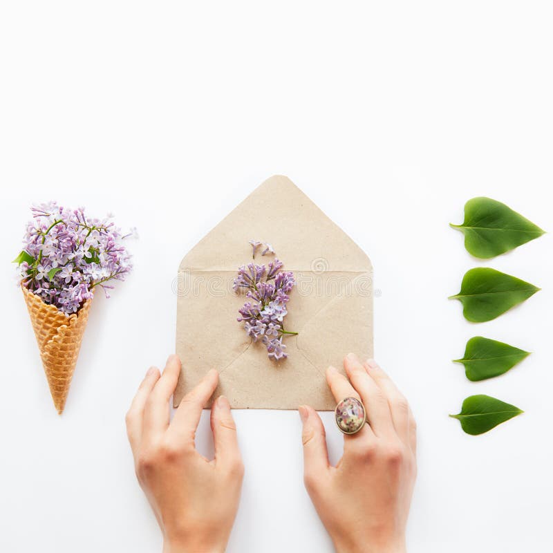 Square card with women hands touching craft paper envelope with lilac flowers surrounded by a waffle corn with a bouquet of lilac