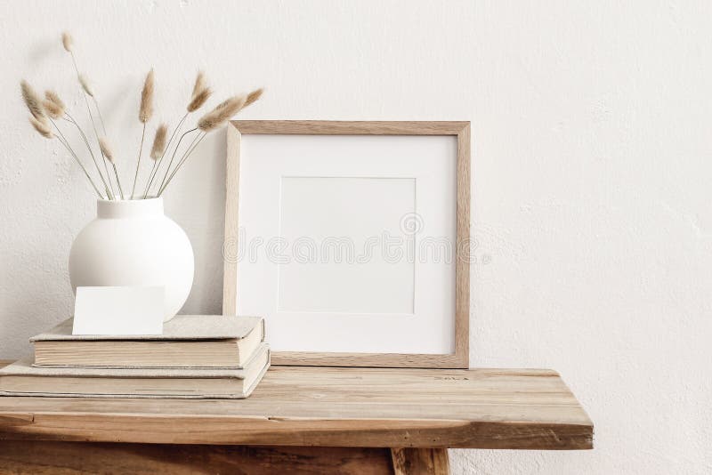 Square wooden frame mockup on vintage bench, table. Modern white ceramic vase with dry Lagurus ovatus grass, books and. Busines card. White wall background