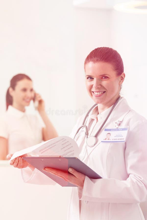 St?ende av att le doktorn som rymmer skrivplattan medan assistentanseende i bakgrund royaltyfria bilder St?ende av att le doktorn som rymmer skrivplattan medan assistentanseende i bakgrund royaltyfria bilder