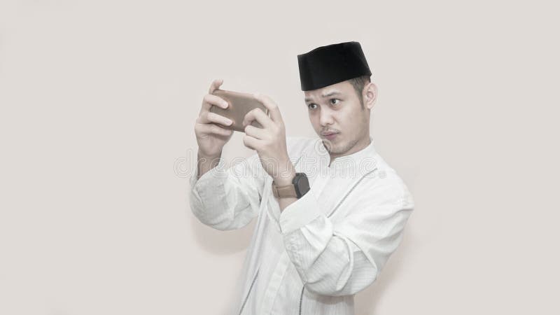 St?ende av den stiliga asiatiska muslim mannen med locket och att anv?nda smartphonen och allvarligt att spela f?r huvud videospe arkivfoto St?ende av den stiliga asiatiska muslim mannen med locket och att anv?nda smartphonen och allvarligt att spela f?r huvud videospe arkivfoto