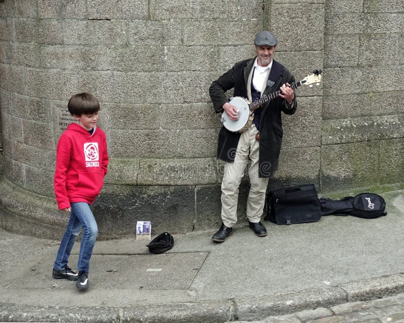 St Ives, Cornwall, UK - April 13 2018: Young boy walking past a busker playing a banjo on a stone walled street corner